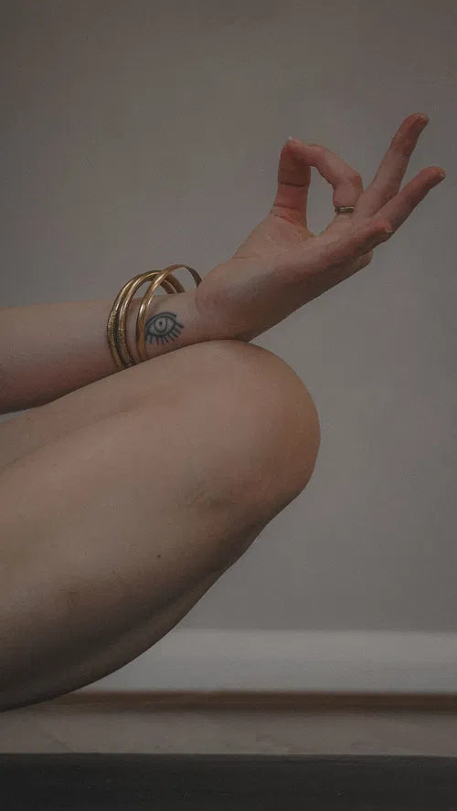 Main posée en mudra sur un genou, avec un tatouage d’œil et des bracelets dorés